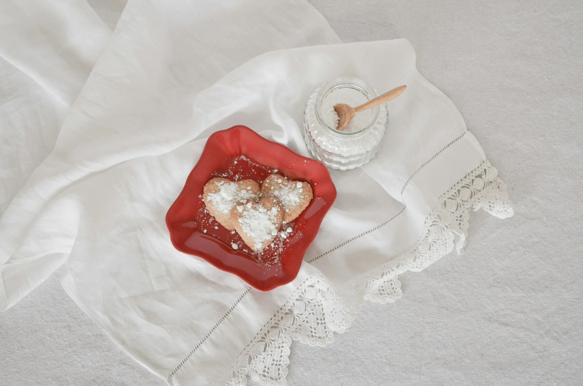 Heart-shaped cookies with powdered sugar on red plate, white linens, glass jar with wooden spoon