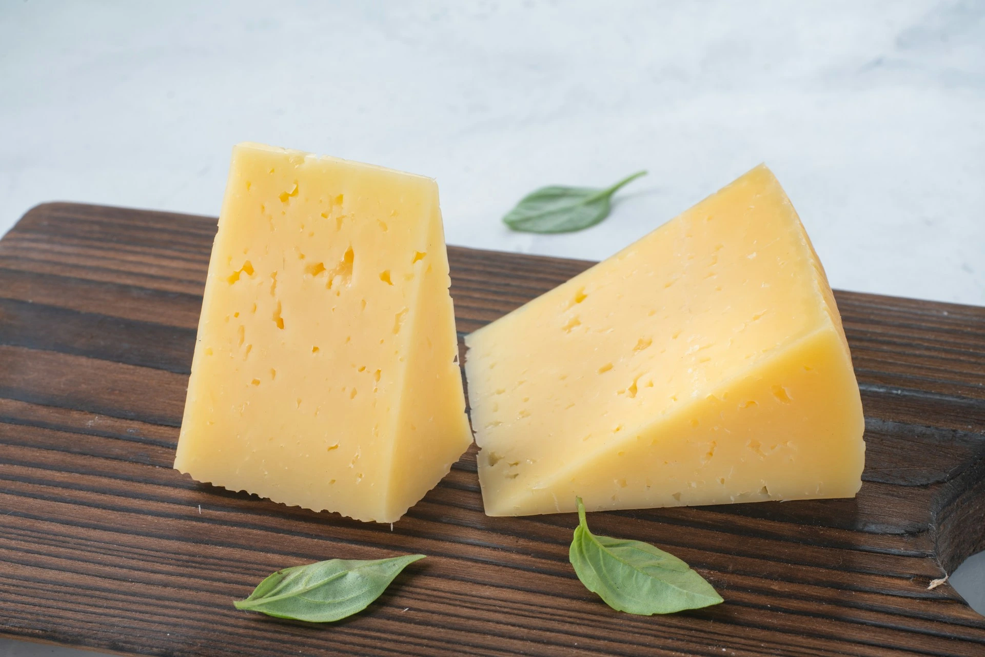 Two wedges of yellow cheese with basil leaves on wooden board