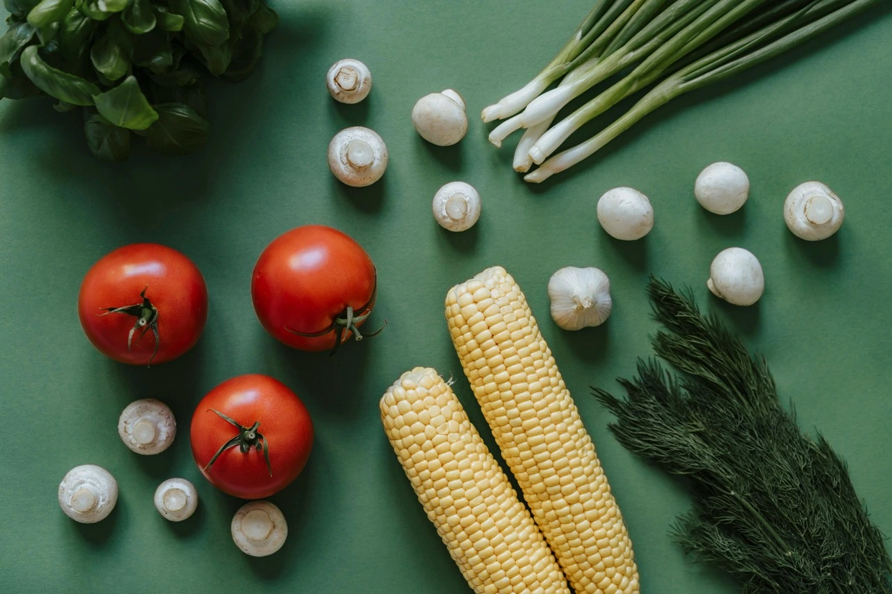 Fresh vegetables arranged on green background: tomatoes, mushrooms, corn, garlic, and green onions.