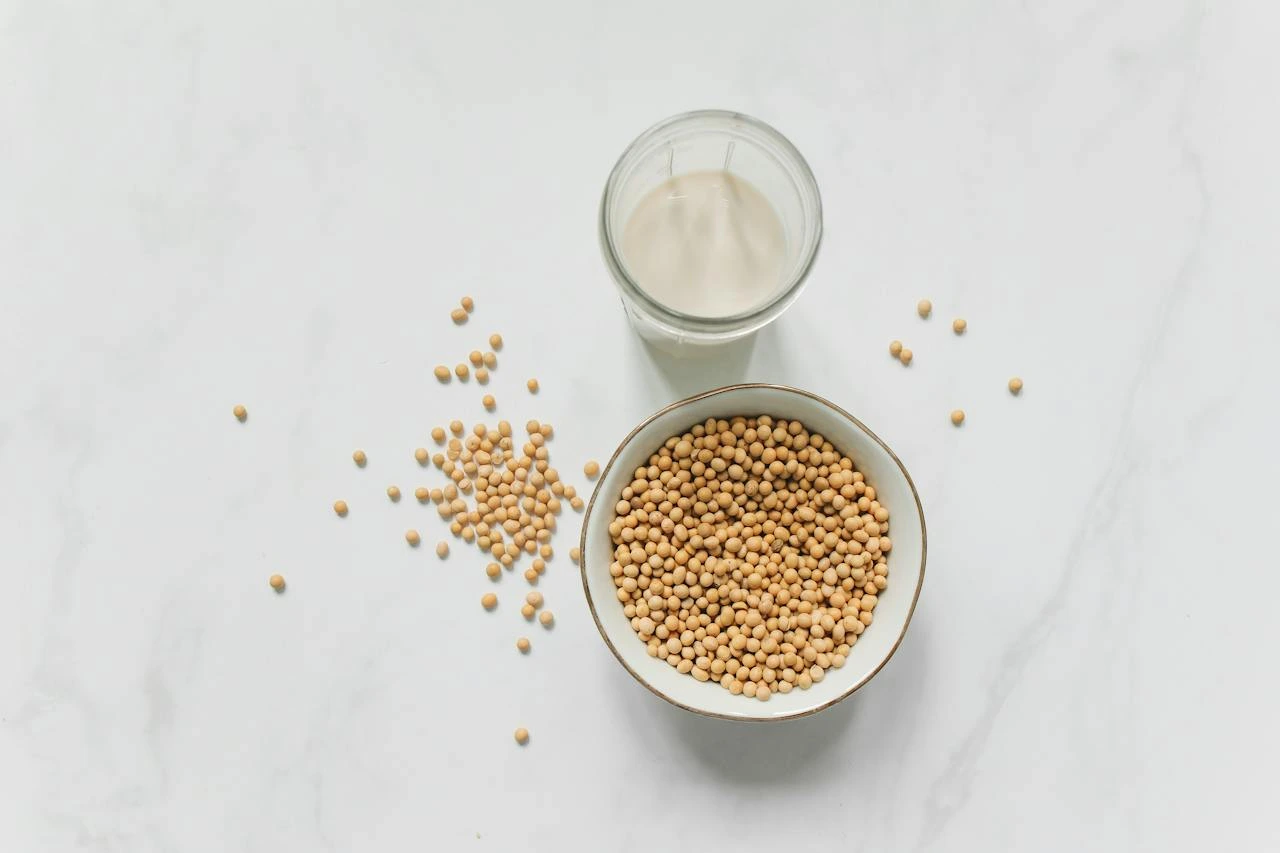 Overhead view of soybeans in a bowl, soy milk in a glass, and scattered beans on white surface.