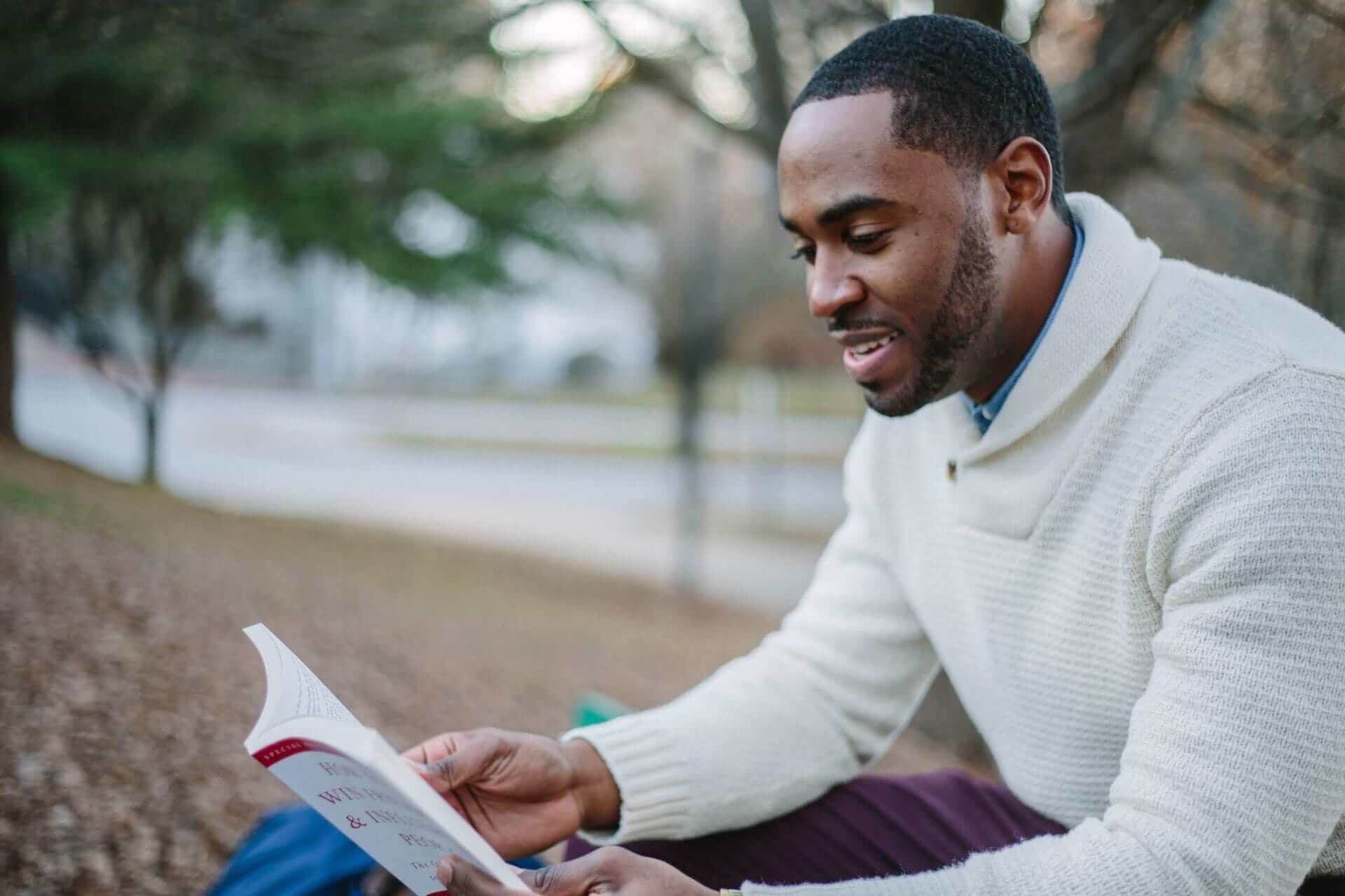 Person reading a book outdoors, wearing a white sweater on a leafy path