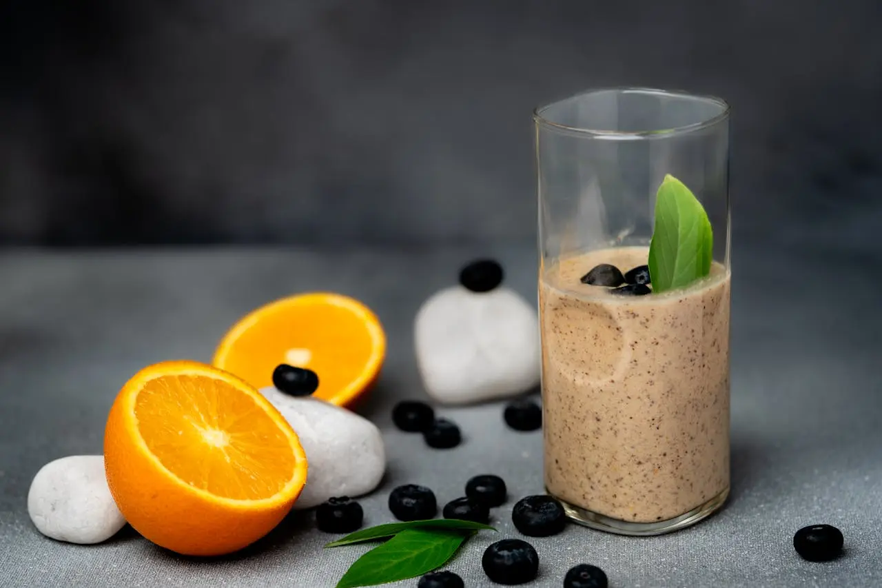 Glass of smoothie with fresh orange slices, blueberries, and mint leaves on gray surface.