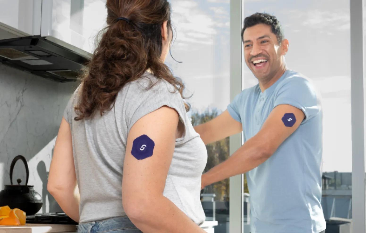 Two people in kitchen, both wearing blue sensor patch, smiling happily