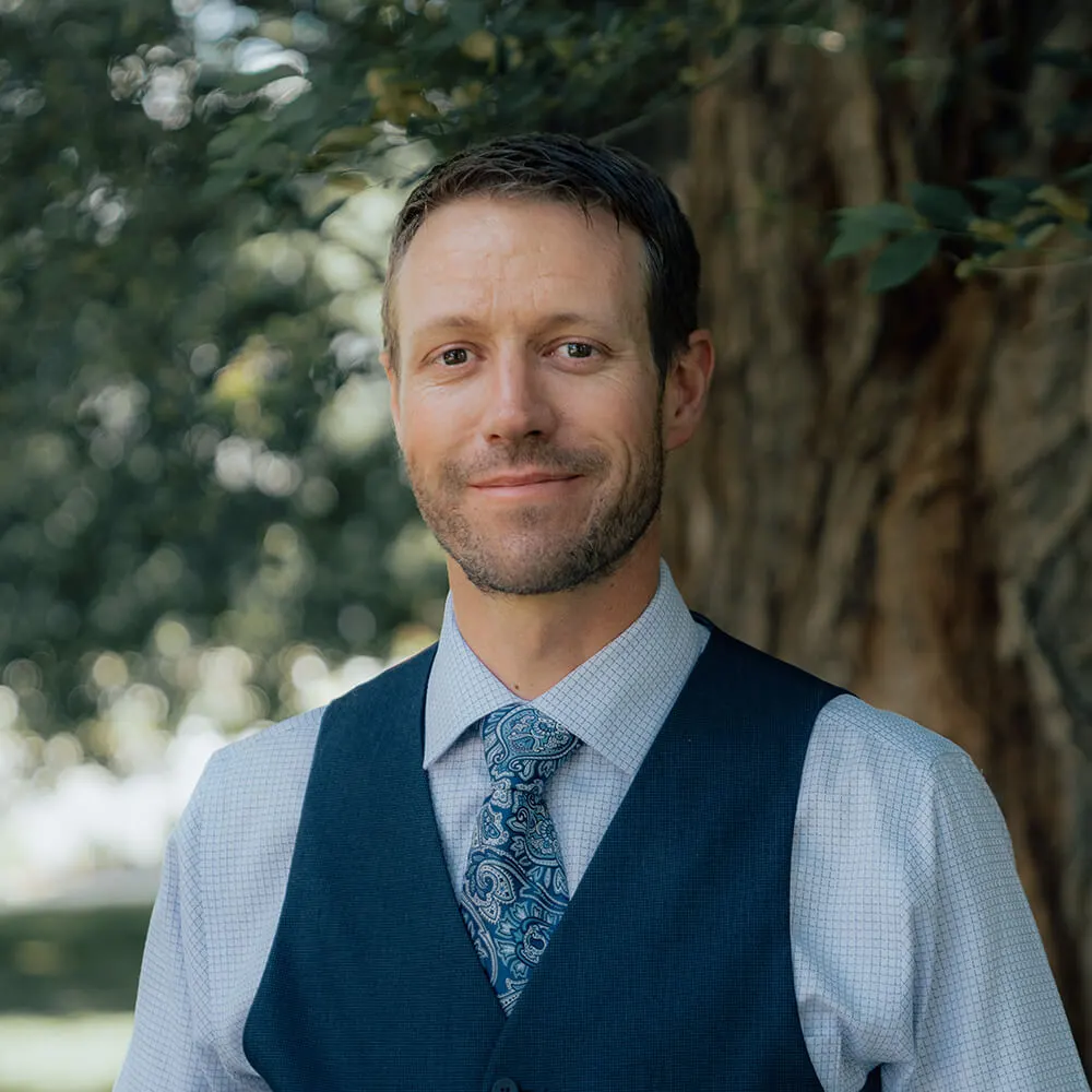 Professional man in navy vest and paisley tie, standing in front of trees