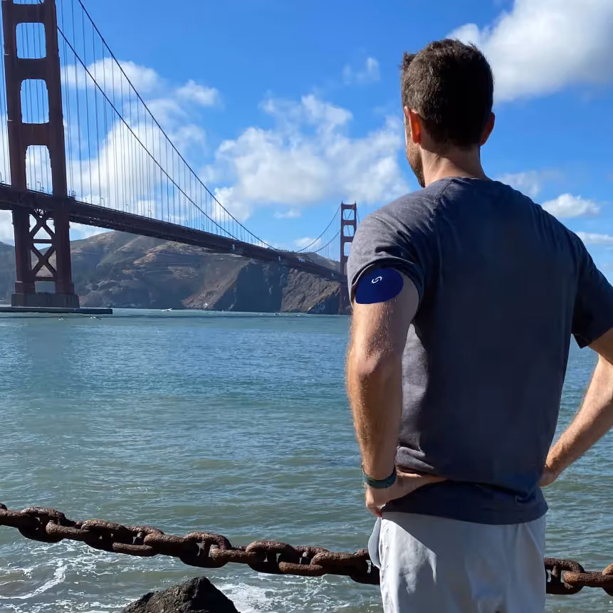 A white man wearing a continuous glucose monitor with a blue Signos sport cover on top on his left arm. His back is facing the camera and he's look at the Golden Gate Bridge.