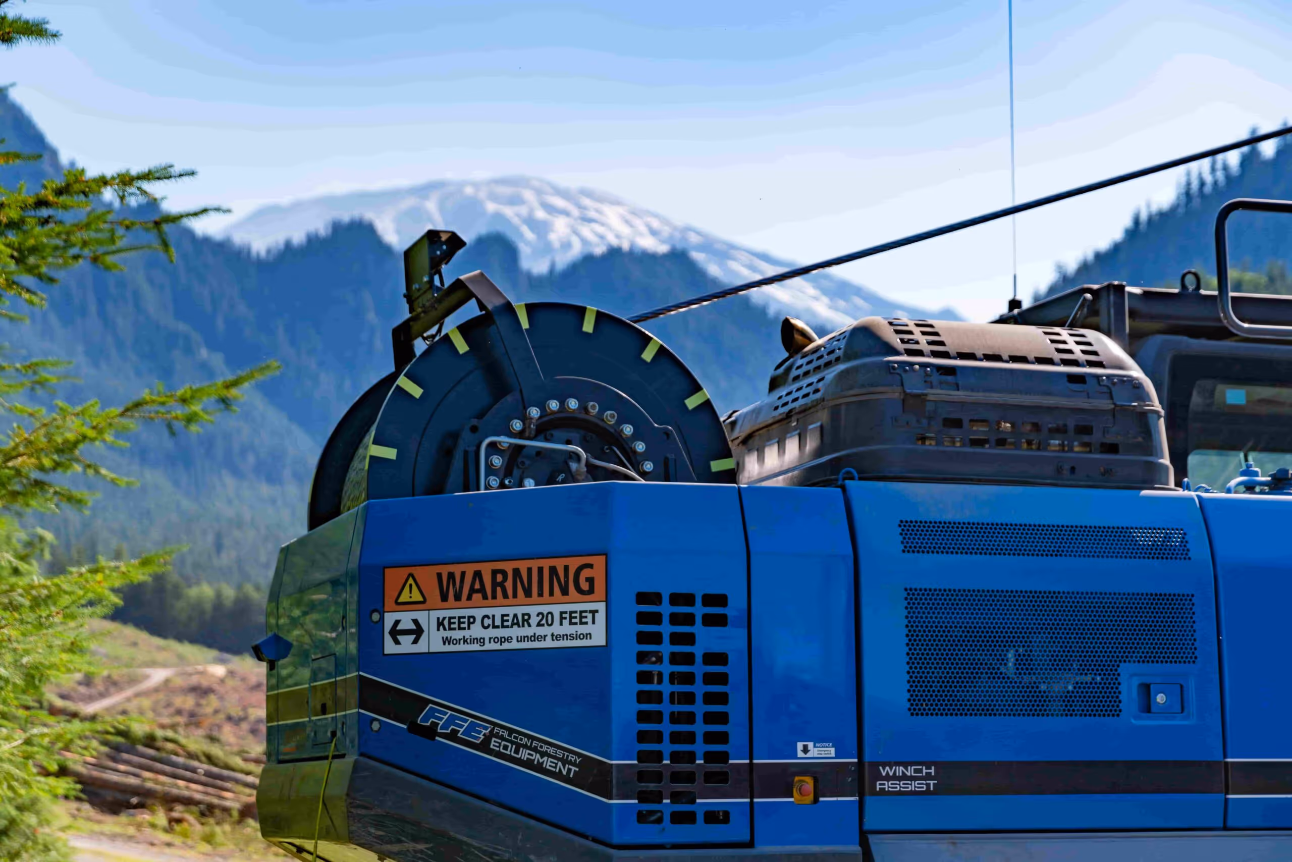 Blue forestry equipment with warning sign and snow-covered mountains in background.