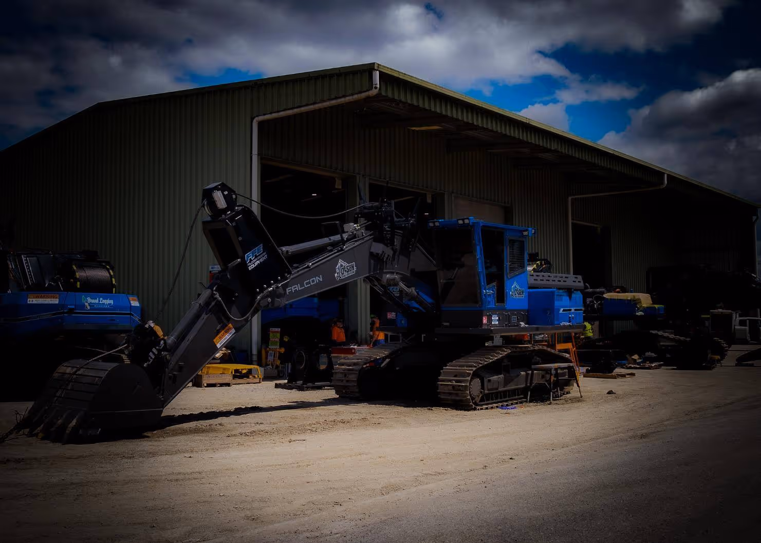 Blue and black tracked excavator parked outside a large metal warehouse under cloudy sky.