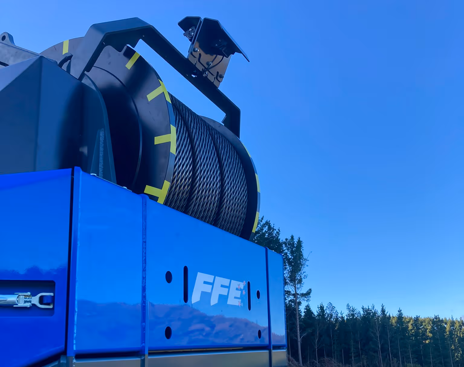 Close-up of a blue industrial winch drum with coiled steel cable and FFE logo against a clear sky.