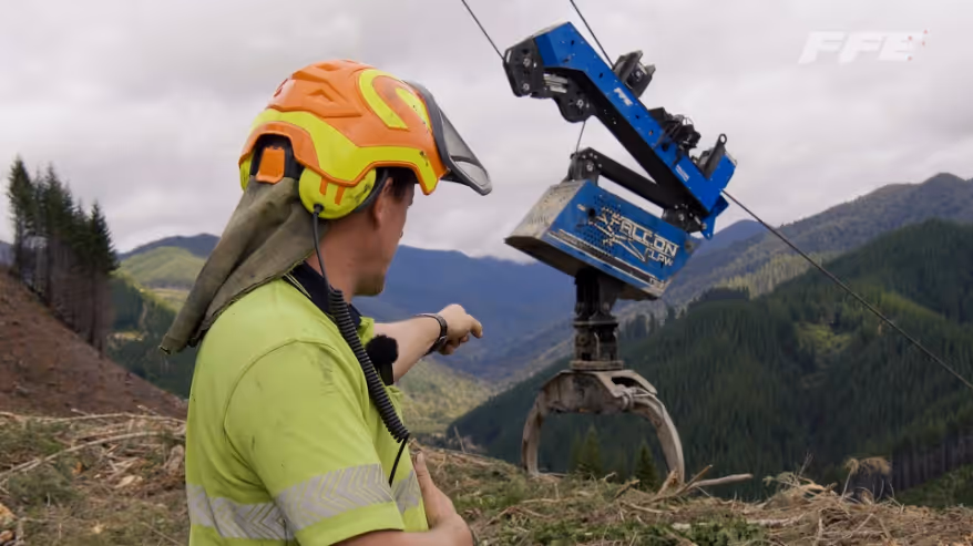 Worker in orange helmet and yellow shirt pointing at blue crane claw in mountainous forest area.