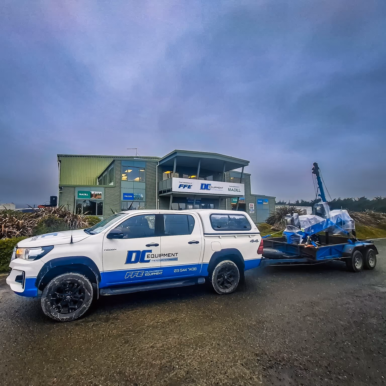 White and blue DC Equipment truck parked outside a green industrial building with a trailer carrying machinery.