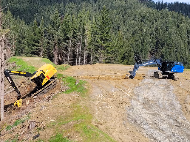 Blue excavator winching up a yellow Tigercat machine on a dirt slope near forest trees.
