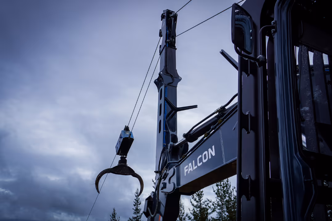 Close-up of a Falcon crane arm with a mechanical claw against cloudy sky and pine trees.