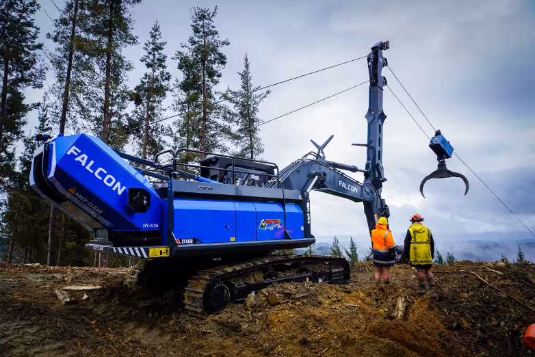 Blue Falcon excavator with claw attachment and two workers in safety gear on muddy ground outdoors.