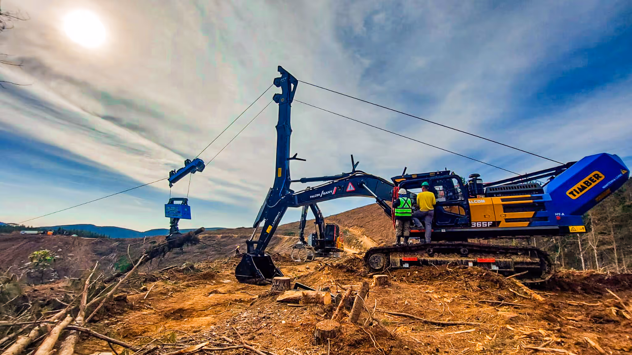 Two workers near large tracked timber harvesting machine lifting logs on cleared land with hills in background.