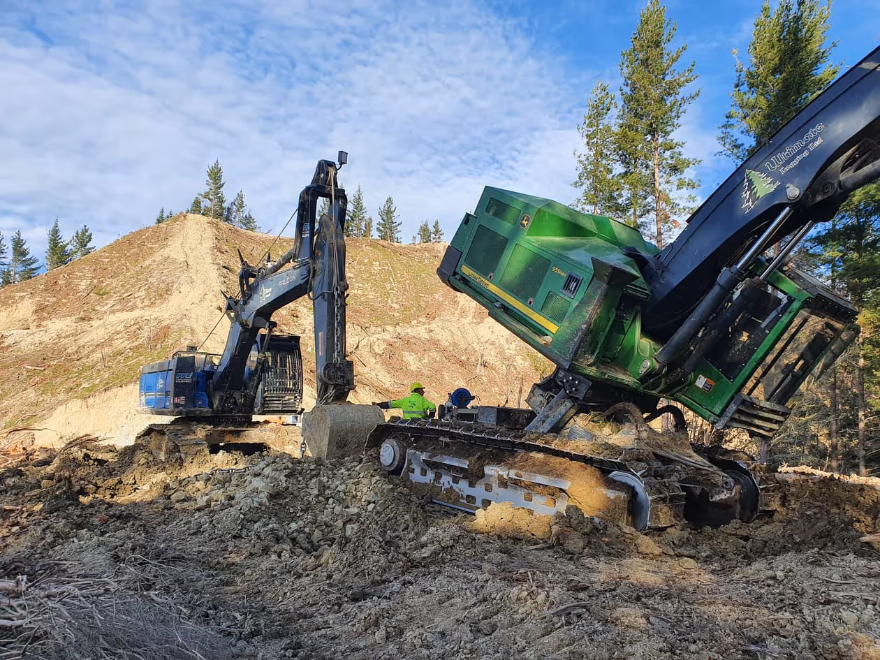 Green tracked excavator stuck at an angle in muddy terrain beside a black excavator and worker.