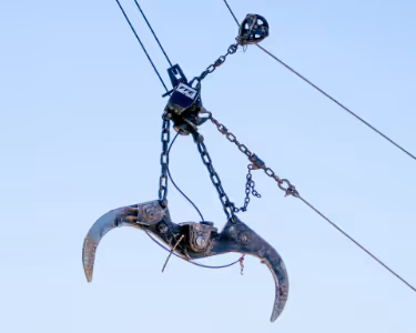 Metal claw crane grapple hook suspended by chains and cables against a clear sky.