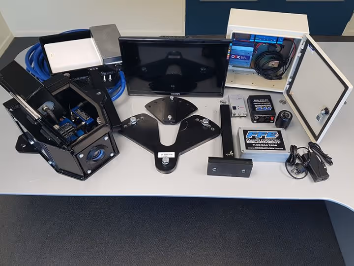 Table with black mechanical parts, wiring, a monitor, and an open white electronics box