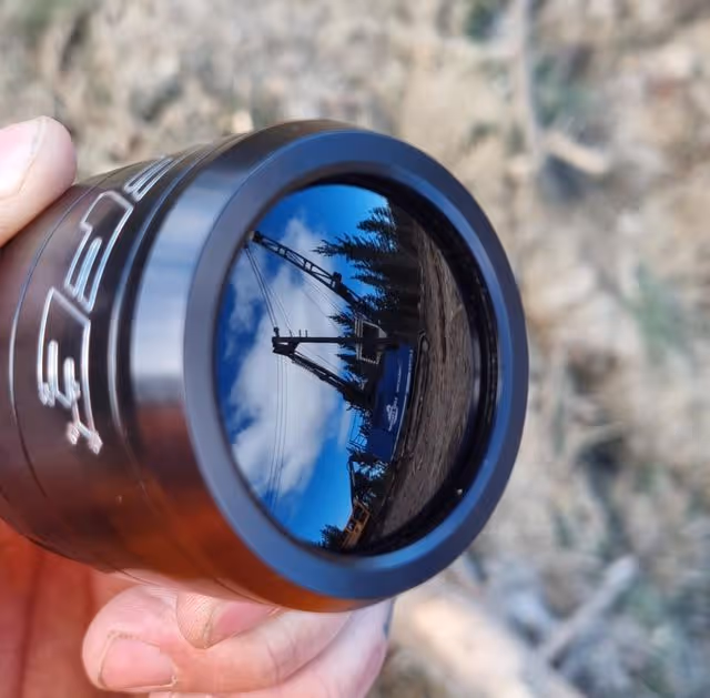 Hand holding black cup reflecting blue sky, trees, and a crane outside on earthy ground.