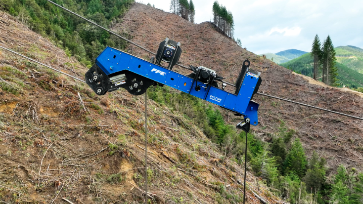 Blue cable carriage suspended on cables over a steep, cleared forest hillside with pine trees.