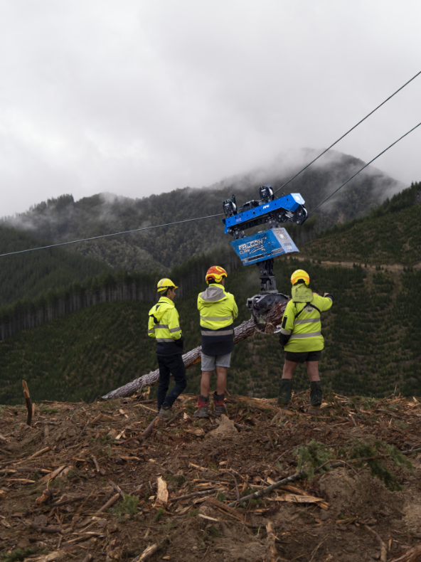 Three workers in helmets and high-visibility jackets watching blue machinery lift a tree trunk on a foggy mountain.