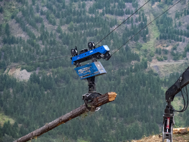 Mechanical harvester gripping a large tree log suspended by cables in a forested mountainous area.