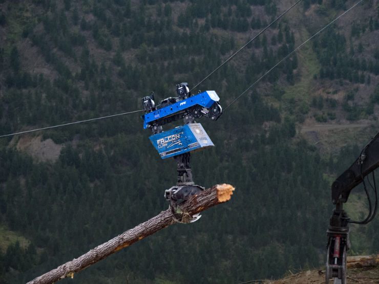 Mechanical grapple attached to cable lifting a large log in a forested mountainous area.