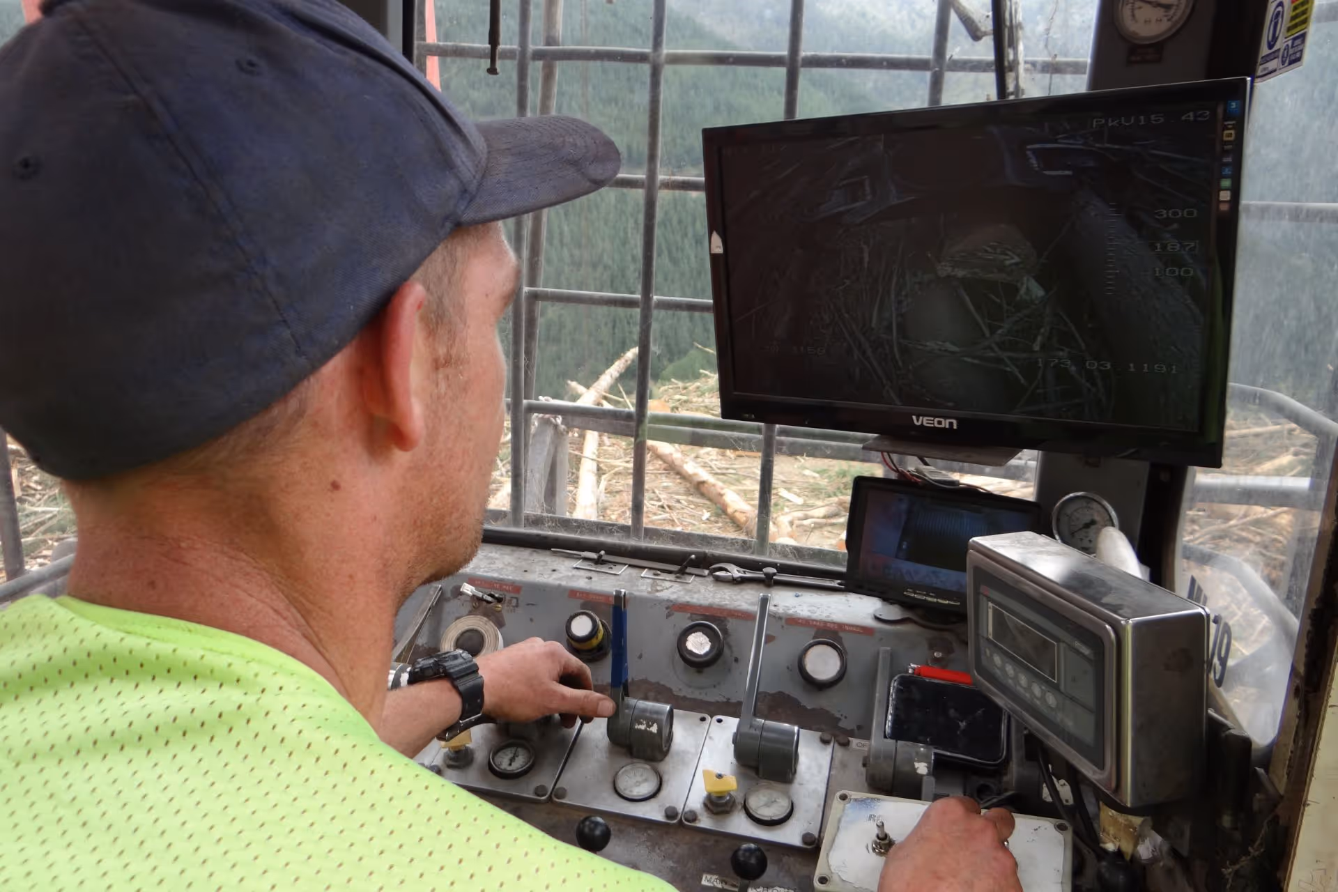 Man operating heavy machinery with control panel and monitors showing outdoor views of logs and forest.