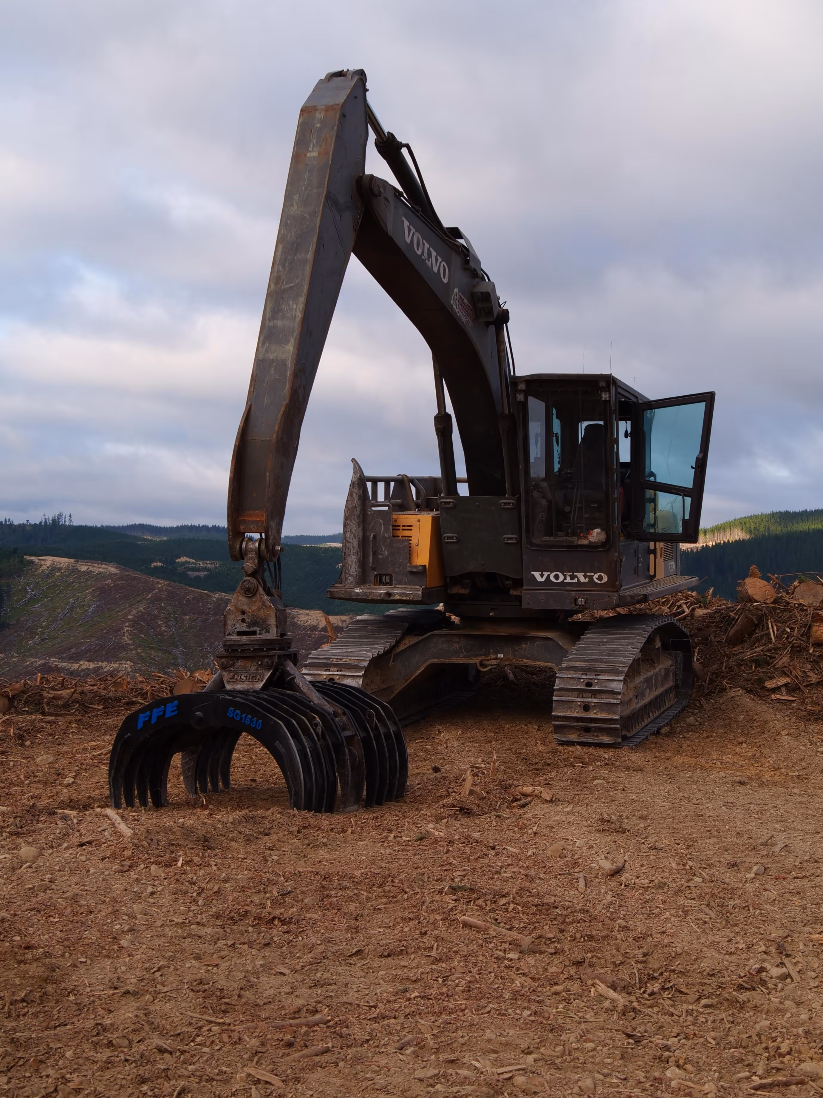 Volvo excavator with claw attachment on dirt clearing near forested hills under cloudy sky.