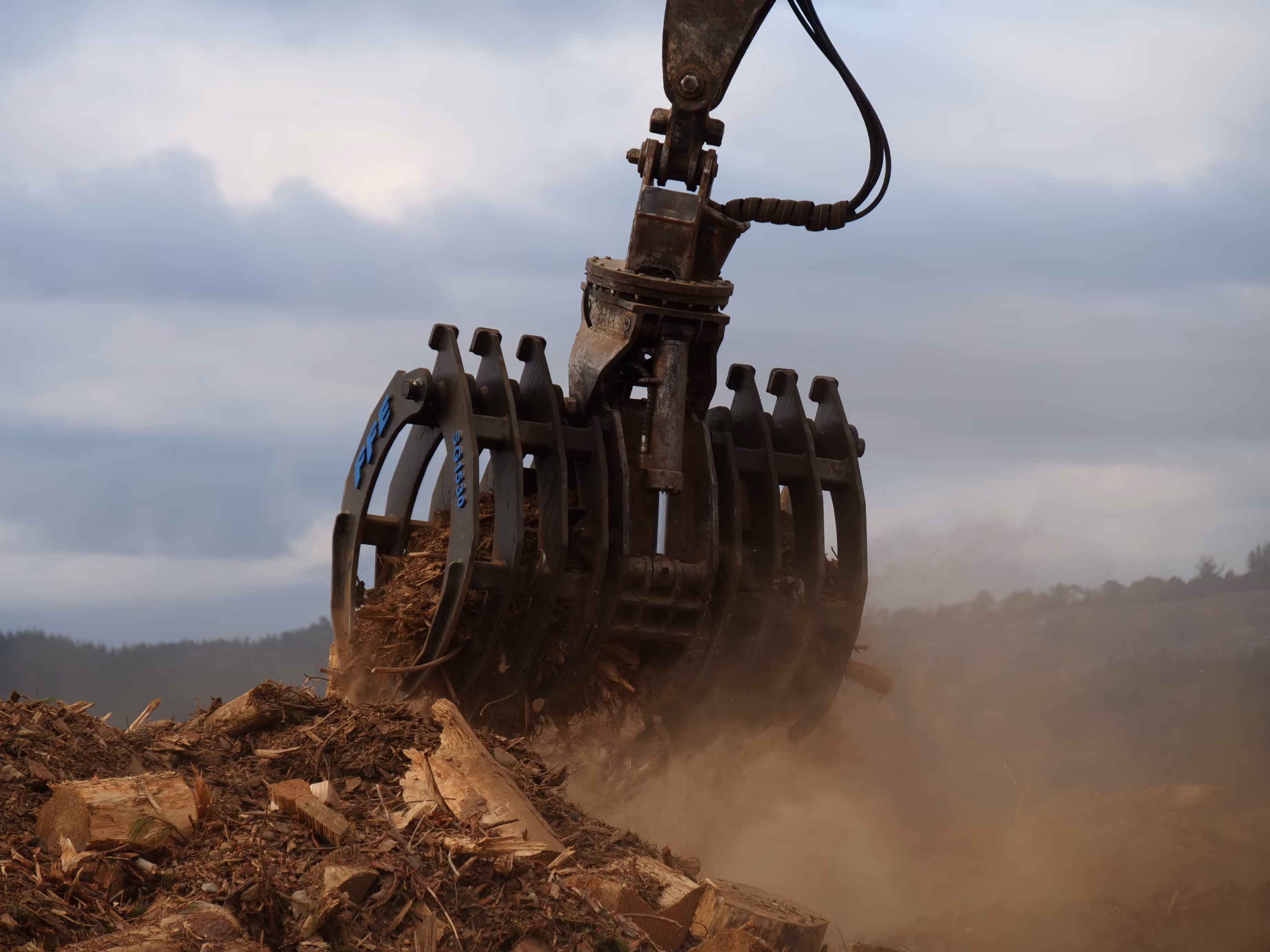 Mechanical claw lifting wood chips and debris with dust around under cloudy sky.