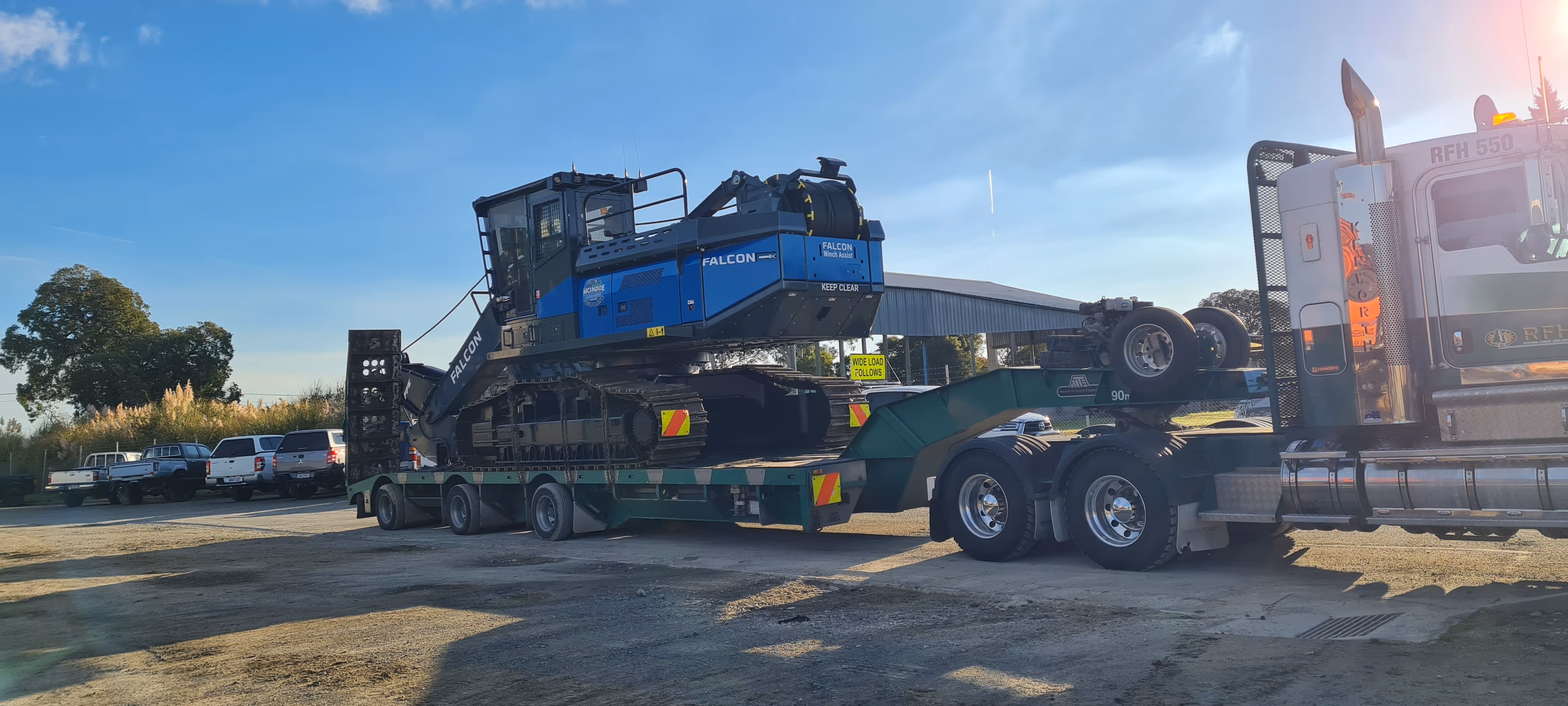 Large tracked excavator labeled Falcon on green flatbed trailer attached to silver semi truck.