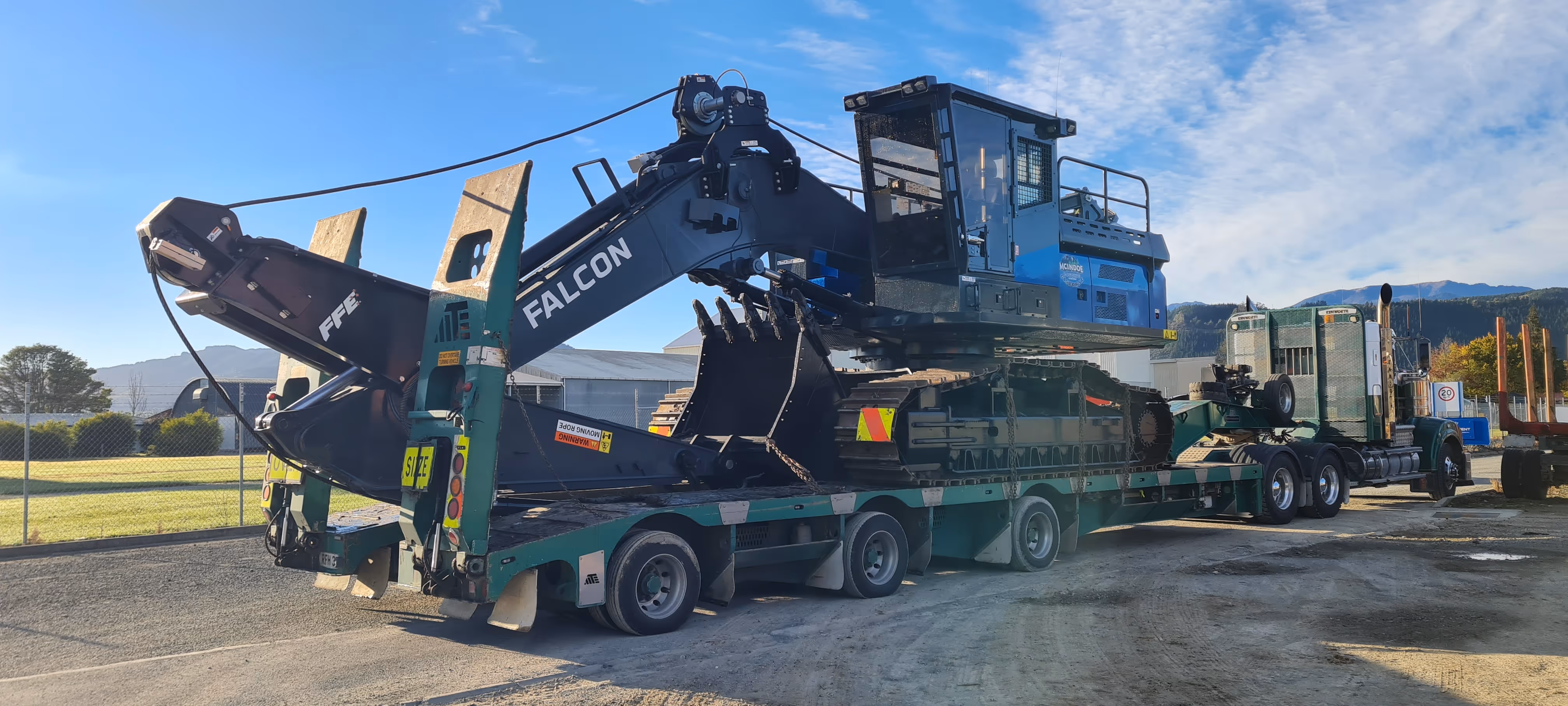 Large blue and black Falcon excavator loaded on green flatbed truck trailer under blue sky.