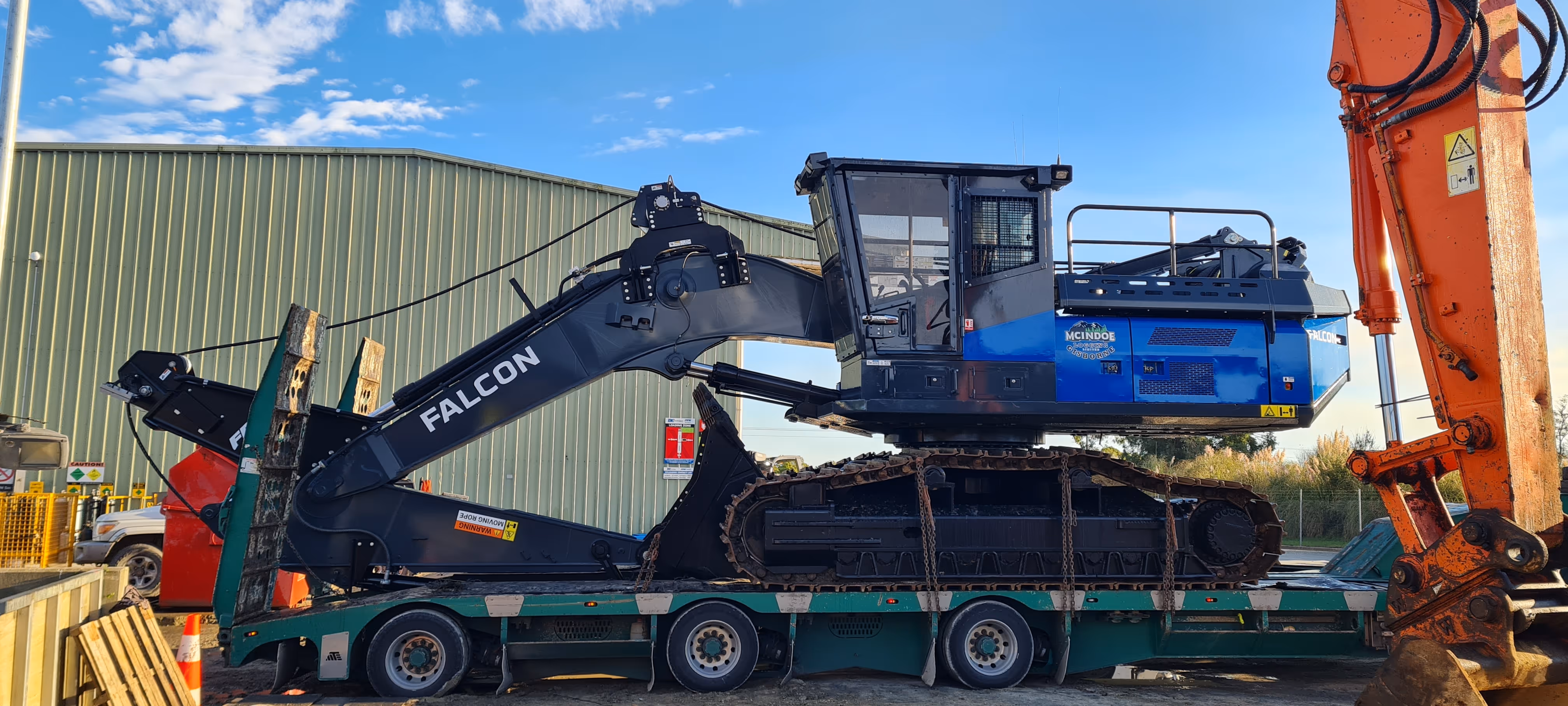 Large blue and black Falcon excavator with tracks loaded on green trailer outside industrial building.