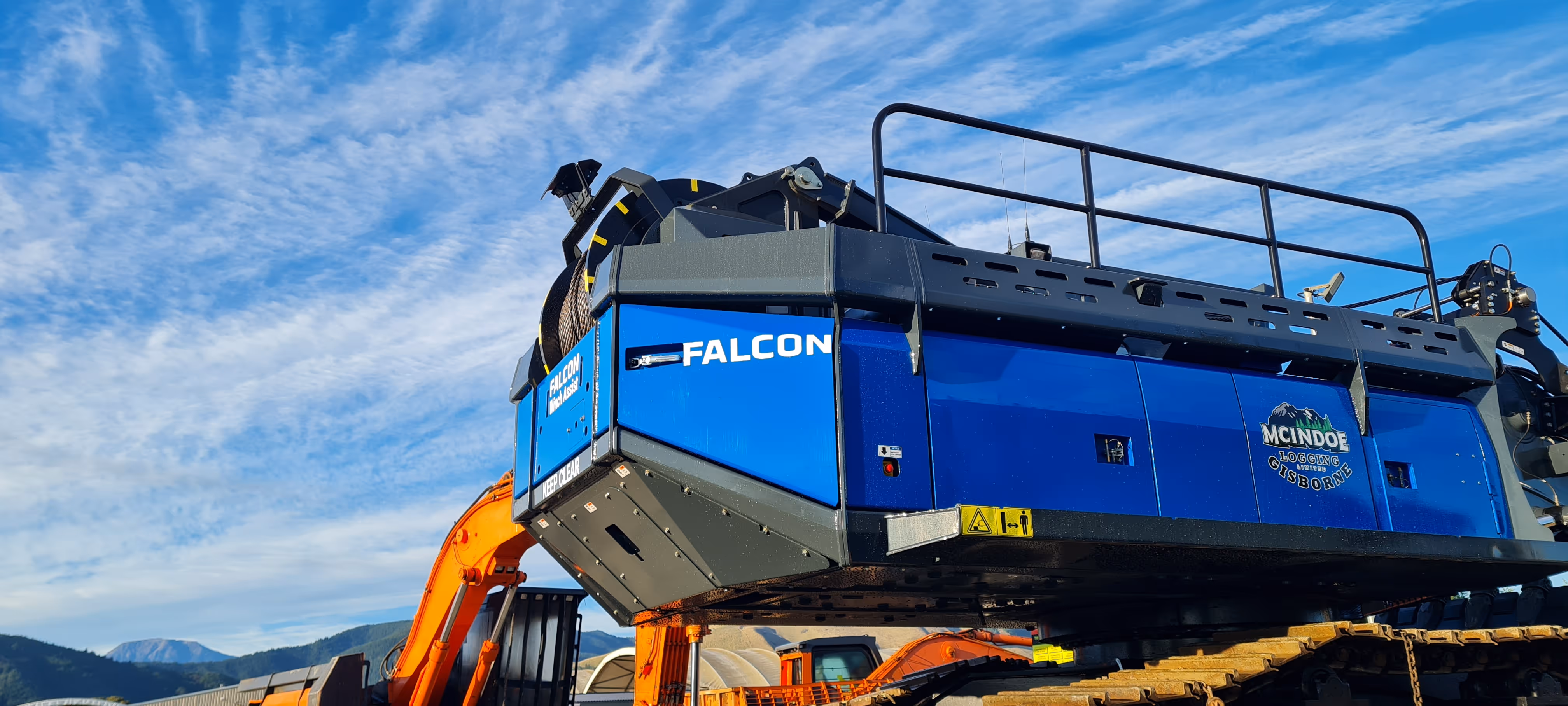 Close-up of blue Falcon McIndoe Logging heavy machinery with an orange arm under a cloudy sky.