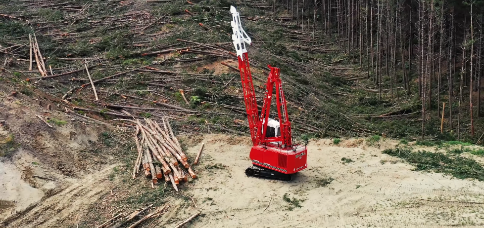 Red swing yarder machine operating in a cleared logging area with stacked logs nearby.
