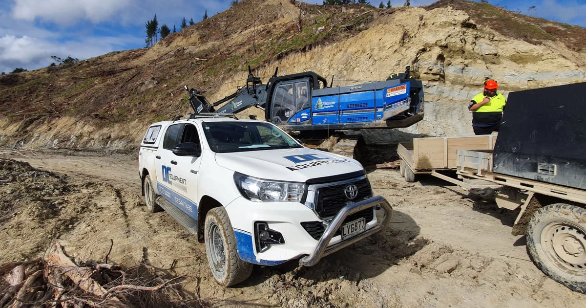 White Toyota pickup with construction equipment on dirt road near rocky hillside and worker in helmet.