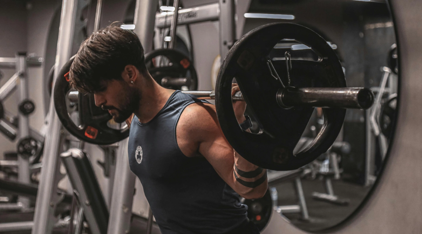 Man with dark hair and beard lifting weights in a gym, wearing a dark tank top. Surrounded by gym equipment, focused expression, dim lighting.