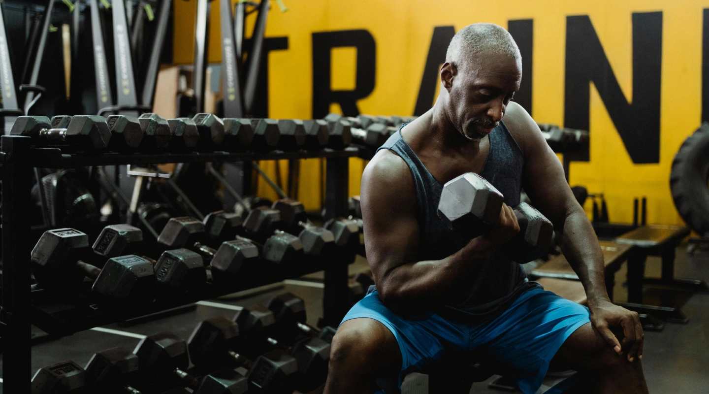 A man in a gym lifts a dumbbell, seated near a rack of weights. He's wearing a sleeveless shirt and shorts, focused and determined. "TRAIN" is visible on the wall.