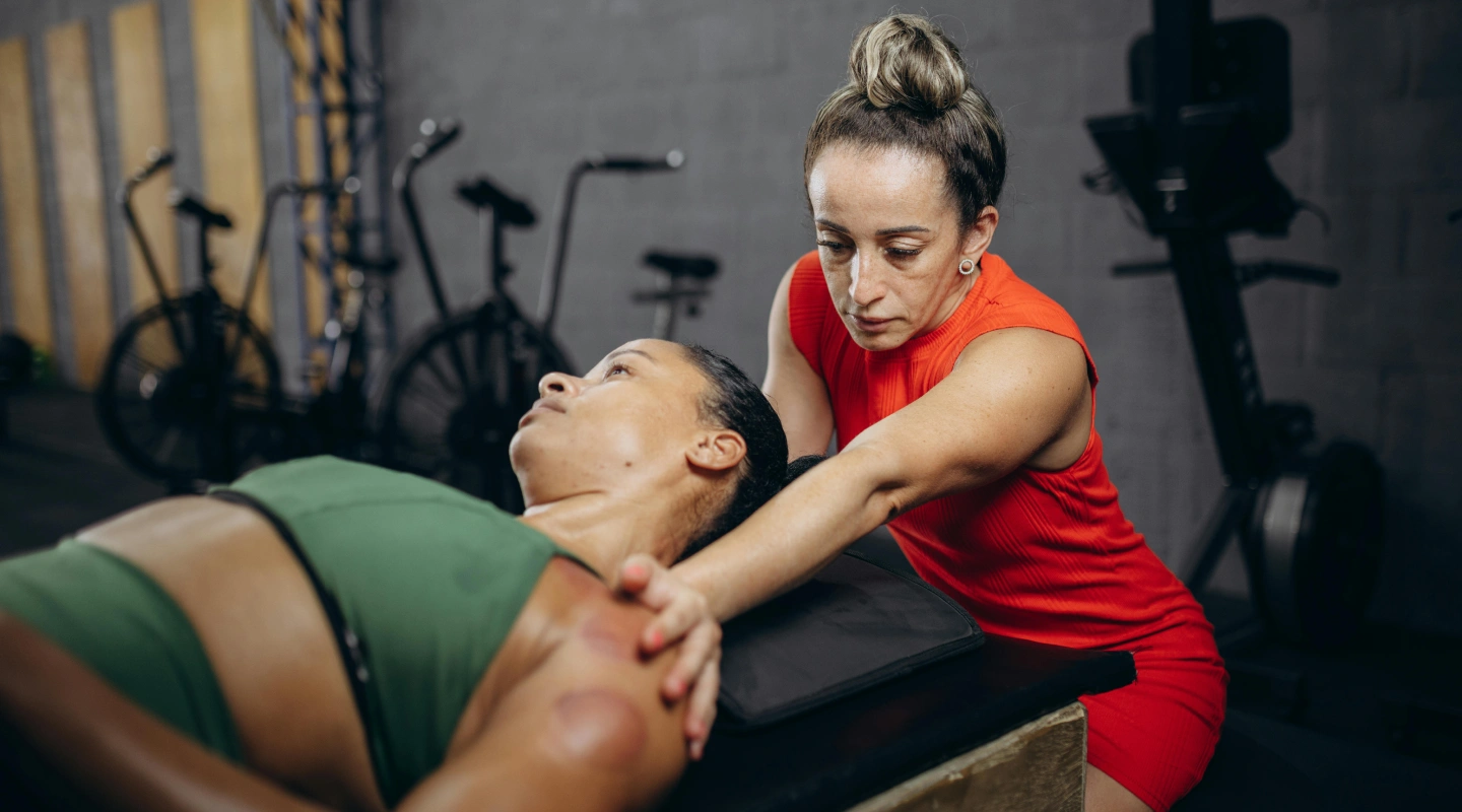 A woman in a red dress attentively stretches another woman's arm on a gym bench. They appear focused, with gym equipment in the background.
