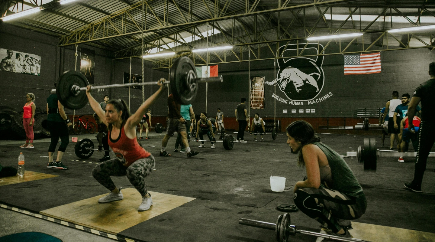 A woman lifts a barbell in a busy gym, surrounded by other focused individuals working out. The atmosphere is energetic and determined.