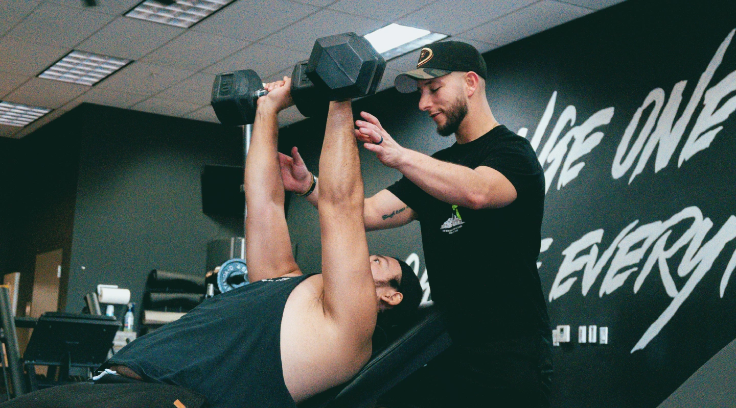 A trainer assists a person lifting dumbbells on a bench in a gym. The trainer offers guidance with a supportive demeanor. Motivational words are visible on the wall.