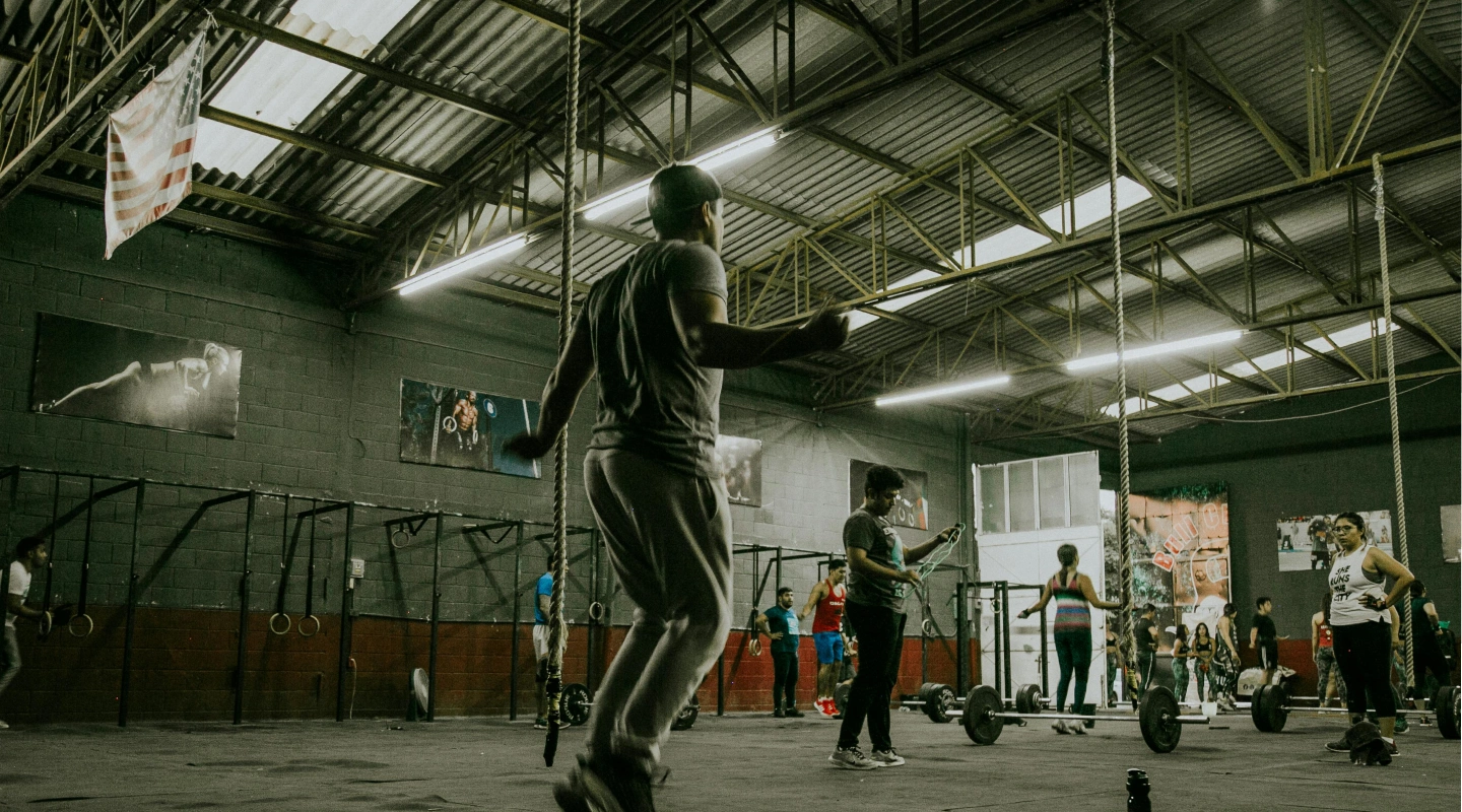 A large gym with people working out; a man skips rope in the foreground while others lift weights. The atmosphere is energetic and focused.