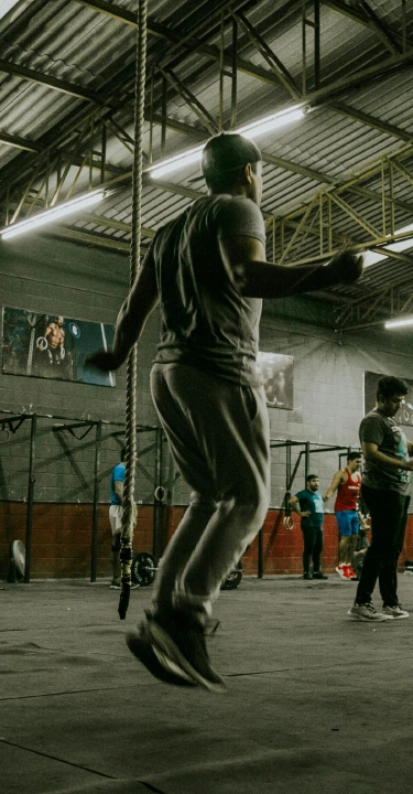 A large gym with people working out; a man skips rope in the foreground while others lift weights. The atmosphere is energetic and focused.