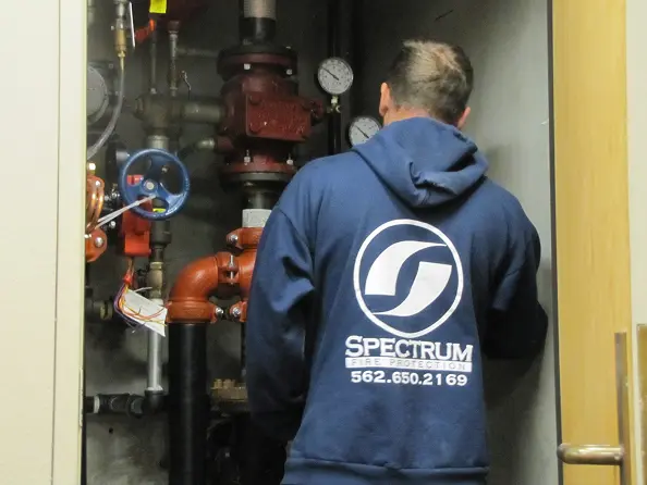 Technician in a Spectrum Fire Protection hoodie working on industrial pipes and gauges in a utility closet.