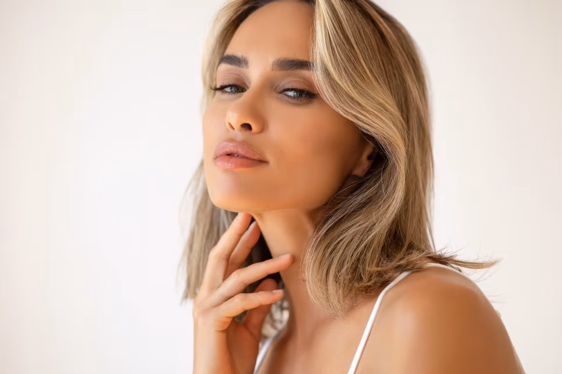 Close-up of a woman with blonde hair and glowing skin gently touching her neck.