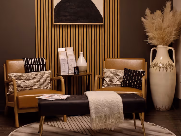 Cozy luxury med spa interior with two tan leather chairs, patterned pillows, a wooden side table, a black bench with a white throw, and a large decorative vase with pampas grass.