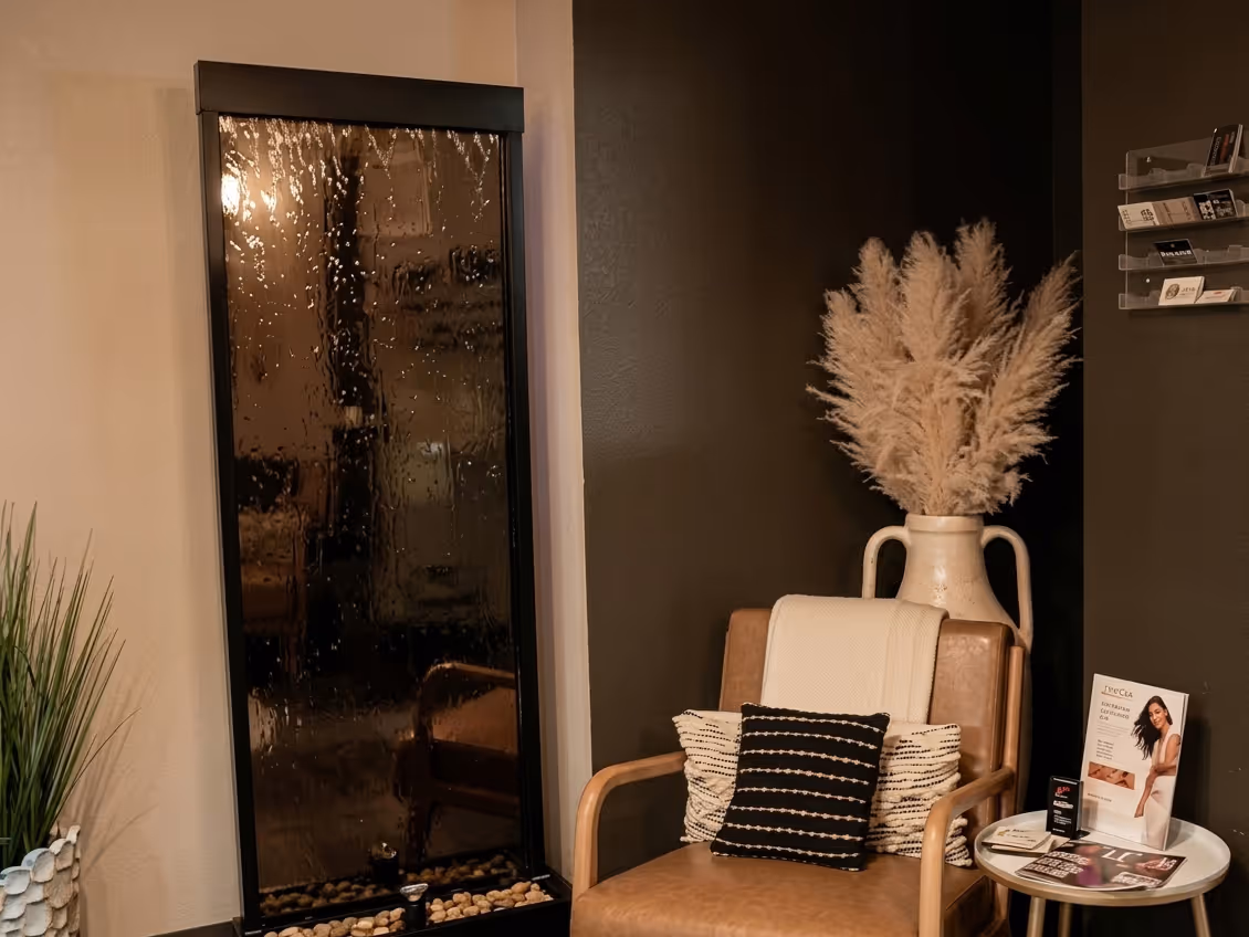 Cozy spa corner with a leather chair, decorative pillows, pampas grass in a vase, and a black water fountain with stones at the base.