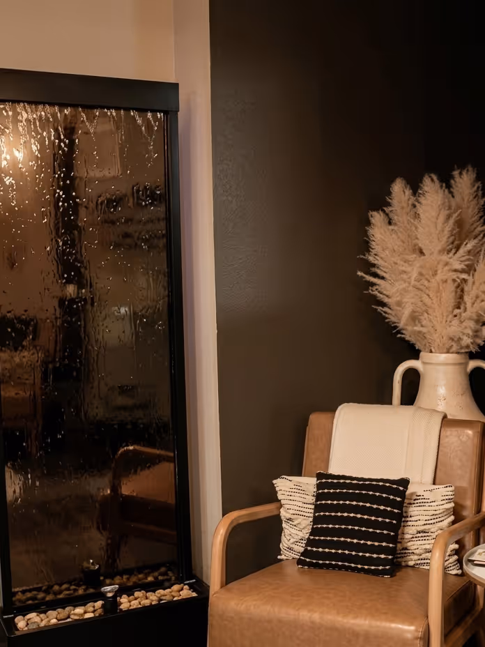 Cozy brown leather armchair with decorative pillows and a cream throw, next to a black indoor water feature and a vase with pampas grass.