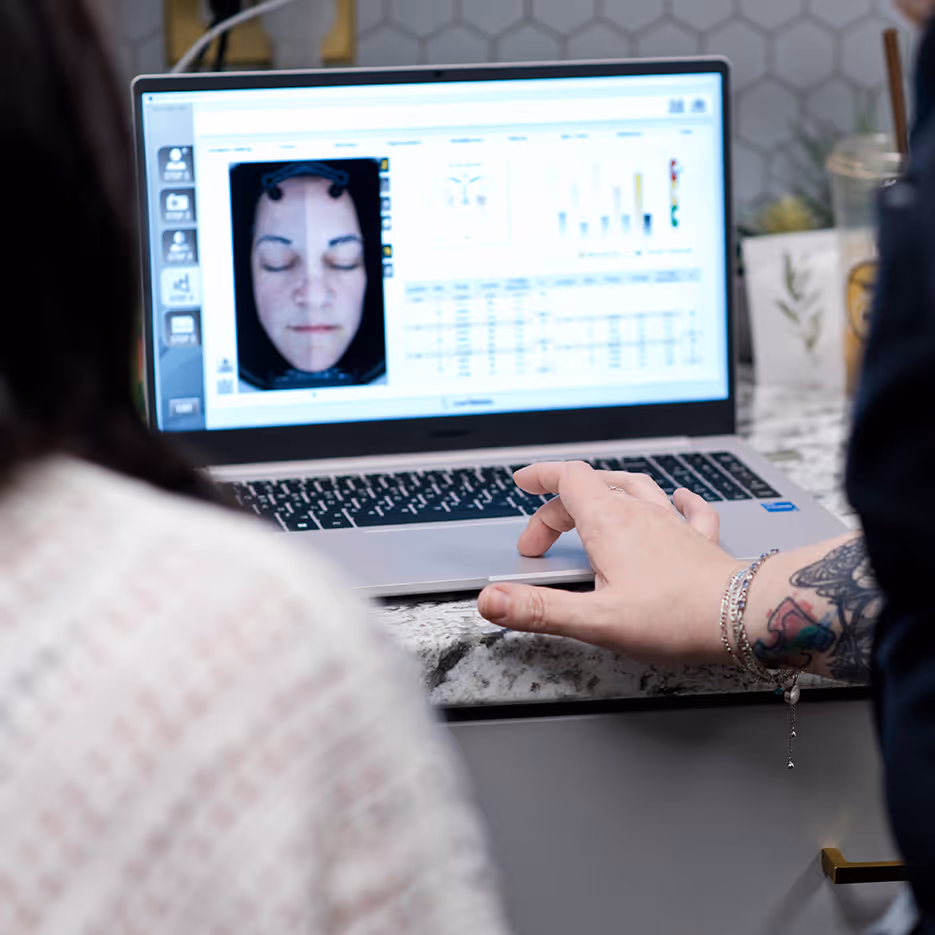 Close-up of a laptop screen showing a facial scan analysis with a person's face and data charts, while a tattooed hand interacts with the keyboard.