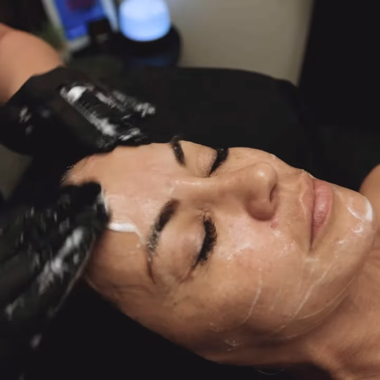 Woman with eyes closed receiving facial treatment with cream applied and gloved hands massaging her forehead.