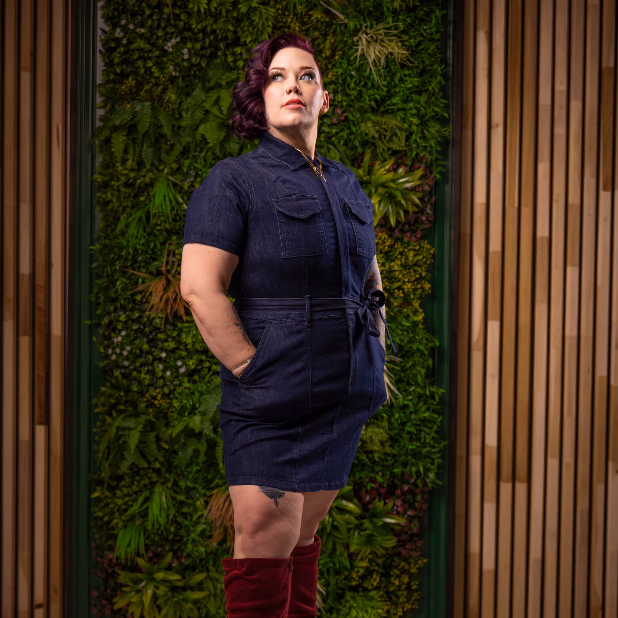 Woman with purple hair wearing a dark blue denim dress and red boots standing in front of a green plant wall and wooden panel backdrop.